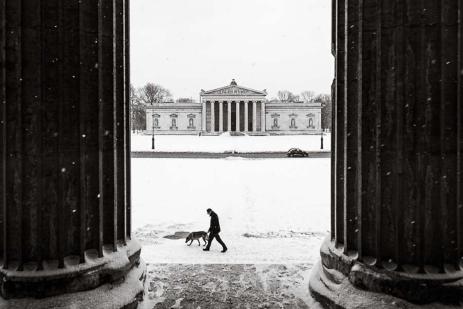 Ein Mann geht mit seinem Hund über den verschneiten Königsplatz, im Hintergrund die Glyptothek.